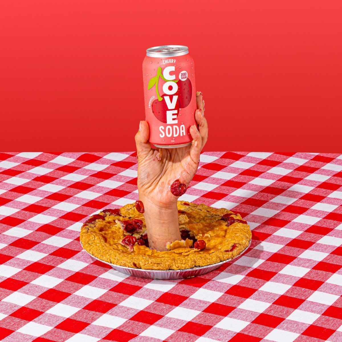 A hand rising from a cherry pie holding a Cove Cherry can over a checkered cloth