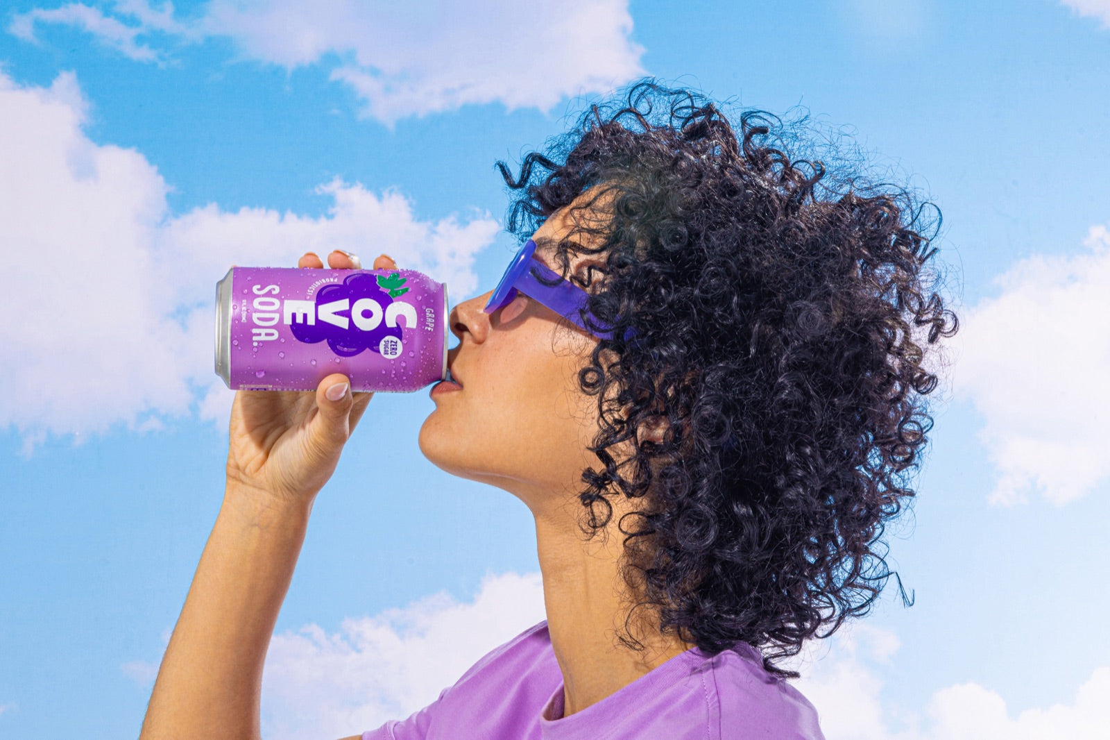 A woman in purple sunglasses sipping a Cove Grape soda against a blue sky