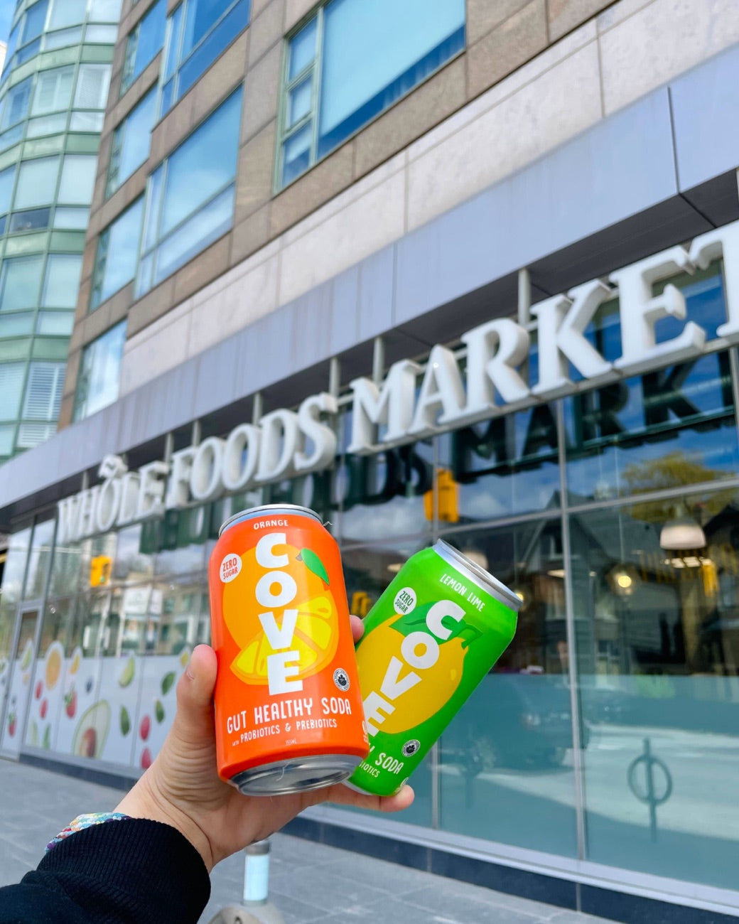 A hand holding two Cove cans up in front of a Whole Foods Market storefront