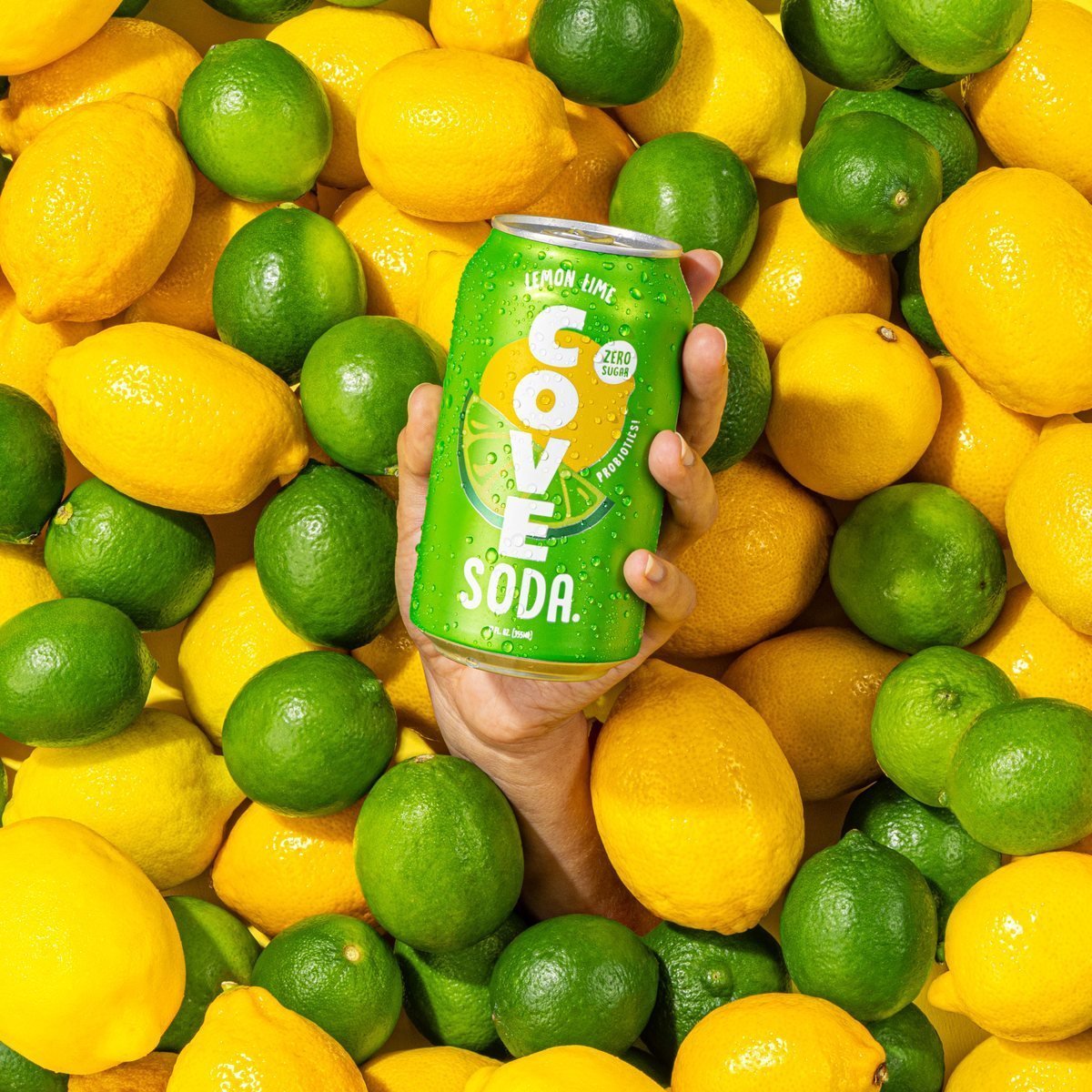 A hand holding a Cove Lemon Lime can among fresh lemons and limes