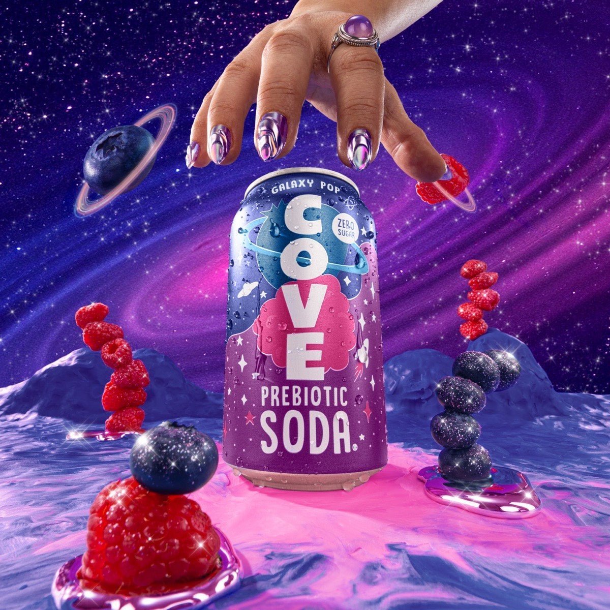 Cove Galaxy Pop can held above a cosmic landscape with raspberries, blueberries and ringed-planet fruit